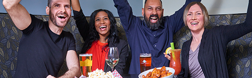 A group of two men and two women sitting in a booth enjoying food and drinks. They are watching a game and cheering.