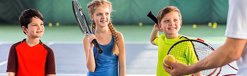 Image of two boys and a girl holding tennis racquets on a tennis court getting instruction from a tennis pro.