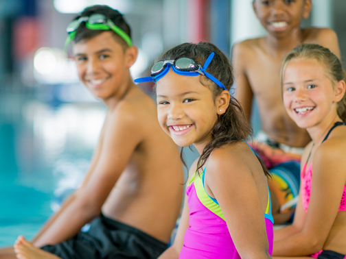 Image of a small group of 8-9 year old's sitting on a pool deck. They are participating in a swim lesson.