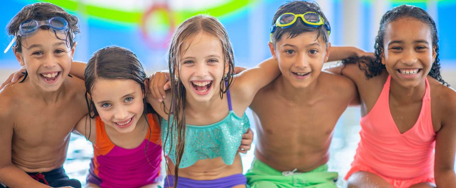Image of a group of kids sitting on the deck of an indoor pool. They have their arms around each other and they are wearing swim suits.