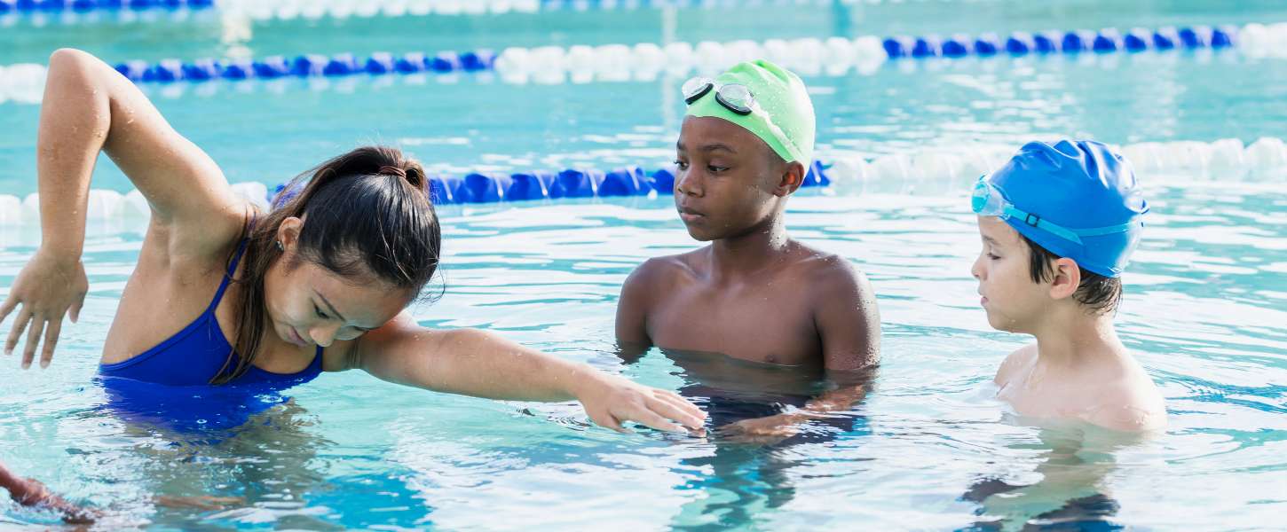 Female swim instructor demonstrating the front crawl to two young boys in an indoor swimming pool.