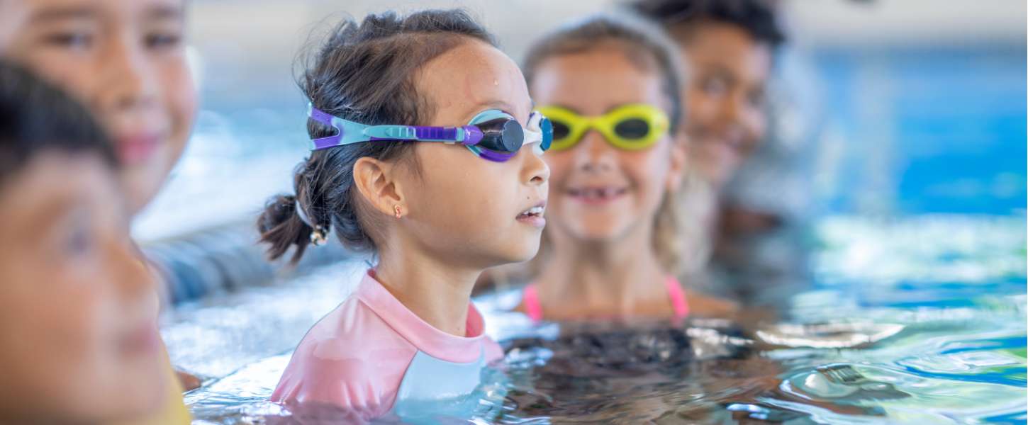 Image of a group of young kids waiting at the side of the pool. They are participating in a swim lesson.