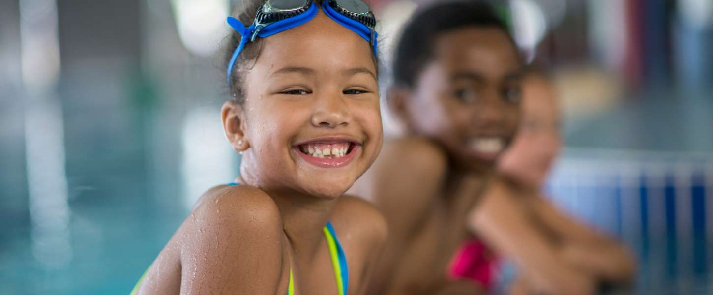 Image of a young girl smiling at the camera. She is wearing a swim suit and having a swim lesson in an indoor pool.