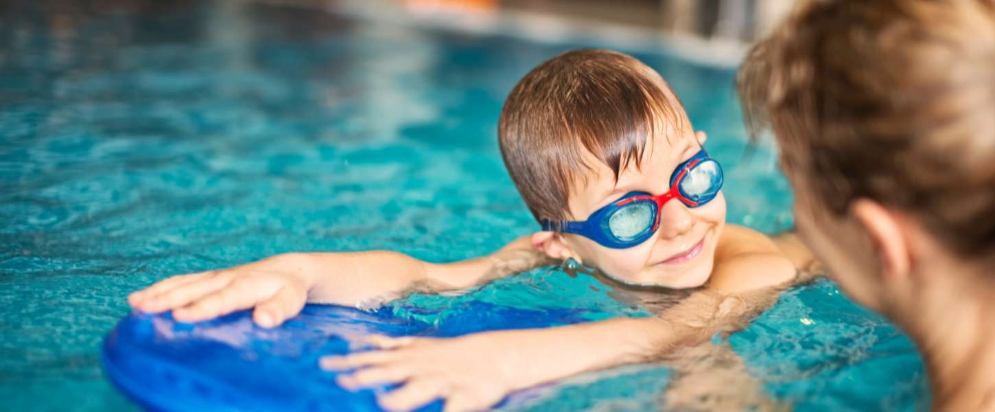 Image of a boy wearing swim googles, holding a flutter board, getting a swim lesson from a female instructor in an indoor pool.