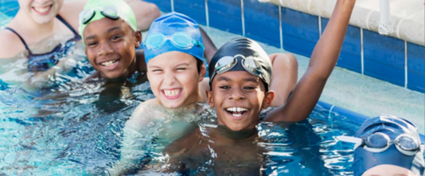 Image of a group of kids in a swimming pool wearing goggles and swim caps getting a swim lesson.